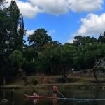 Scenic view of Kastel Fortress in Banja Luka with people riding a traditional Dayak boat on the Vrbas River and others fishing along the riverbank, highlighting a blend of history, culture, and leisure.